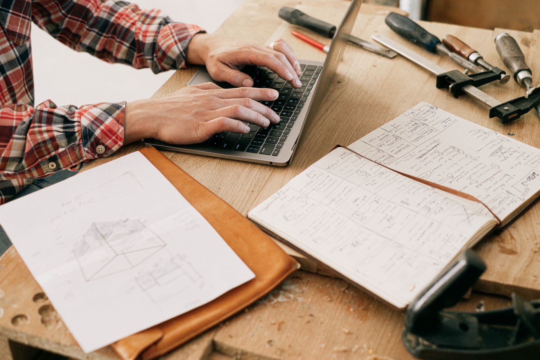 Hands typing on a laptop at a workbench, with tools and a sketchbook beside it