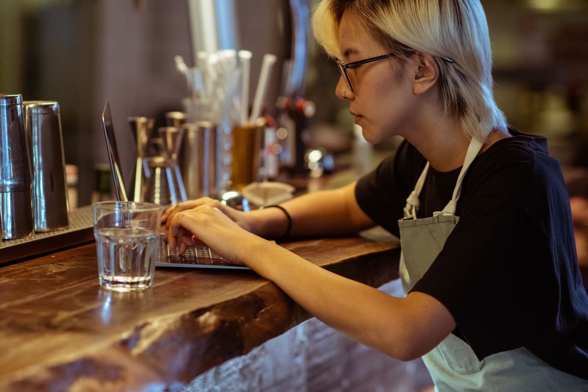 A café worker in an apron typing on a laptop at a wooden bar counter