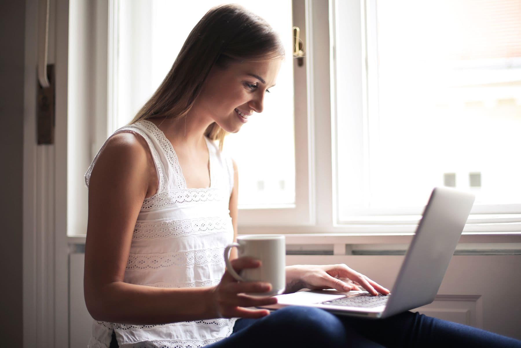 A woman by a window holding a mug and typing on a laptop